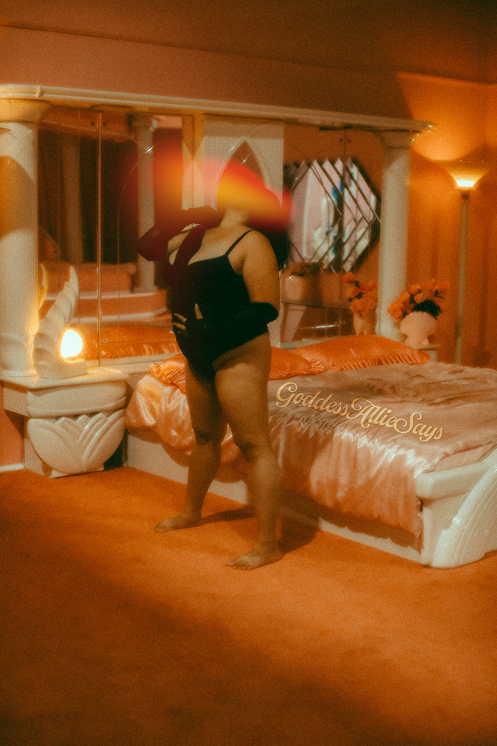 Allie stands in front of a bed wearing a black bodysuit holding a black flogger, her head leaning back and her stance wide.